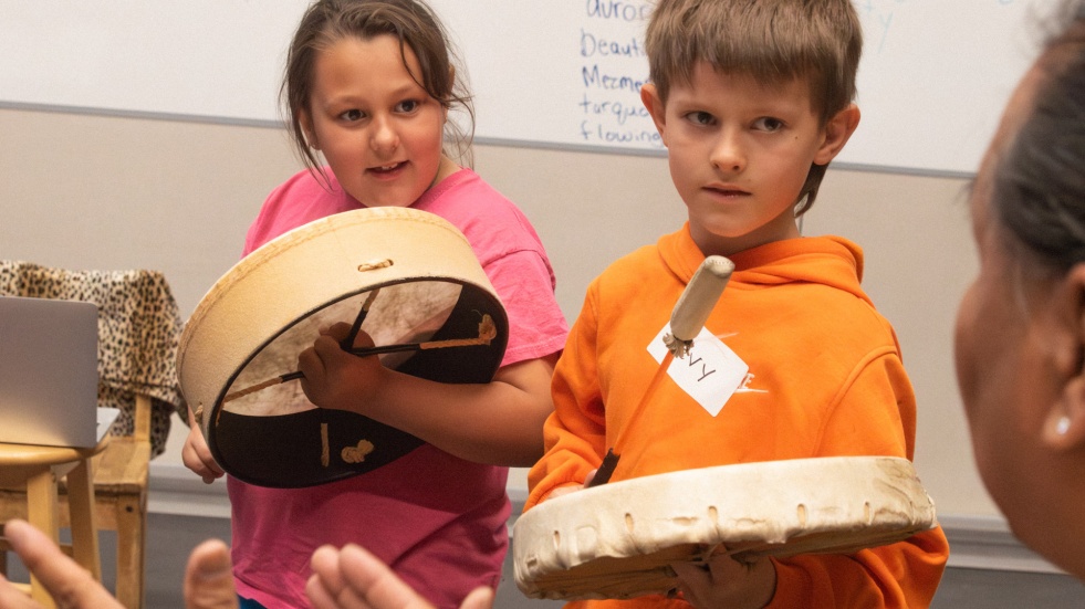A young boy and girl hold hand drums and watch an instructor 
