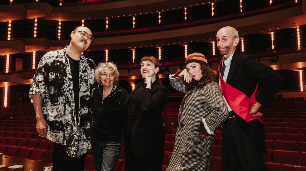 A group of five people stand together on a theatre stage, posing in front of rows of red seats and illuminated balconies. They are dressed in a variety of outfits, including patterned jackets, a long coat, and a sash. The lighting emphasizes the larg