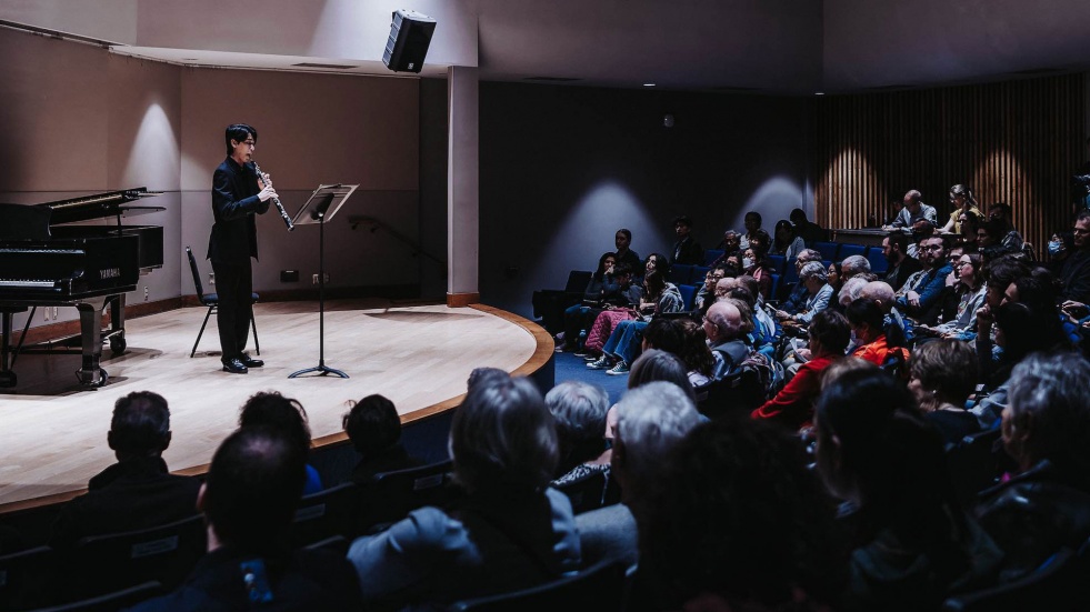 An oboist performs on stage in a small concert hall while a seated audience watches attentively.