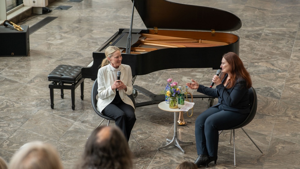 Two people talking in front of a grad piano, with an audience watching.