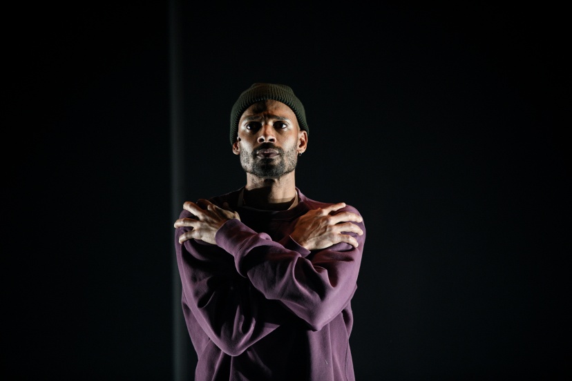 Performer wearing a beanie with arms crossed over their chest, illuminated on a dark stage