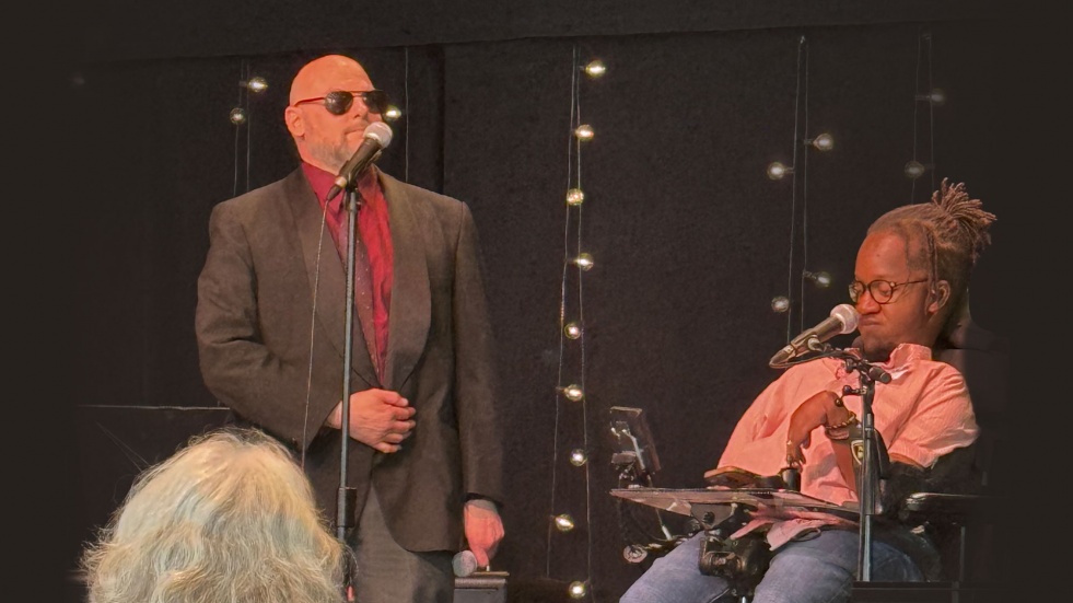 Two speakers on a stage at an accessibility-focused event. One speaker stands at a microphone while another speaker uses a power wheelchair and speaks into a microphone. String lights hang against a dark backdrop, and audience members are seated in t