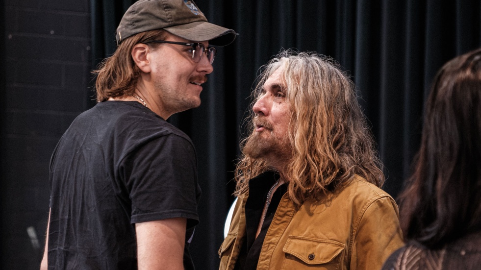 Two men stand face to face in conversation during a rehearsal, one in a ball cap and glasses, the other with long grey hair and a tan jacket.