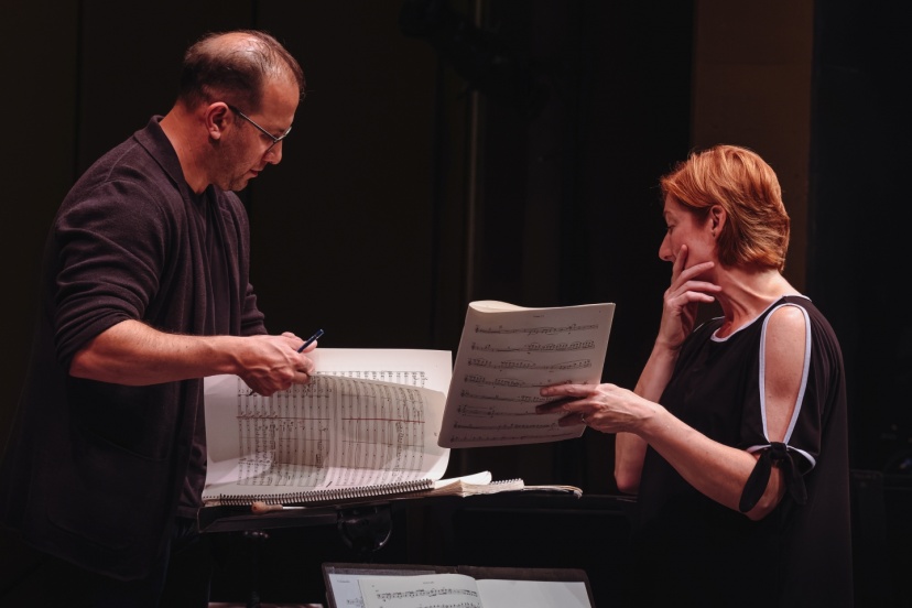 A musician and conductor stand at a music stand onstage, reviewing printed scores together. One page of a large orchestral score is turned while the other person studies a separate sheet of music. Warm stage lighting illuminates the scene against a d